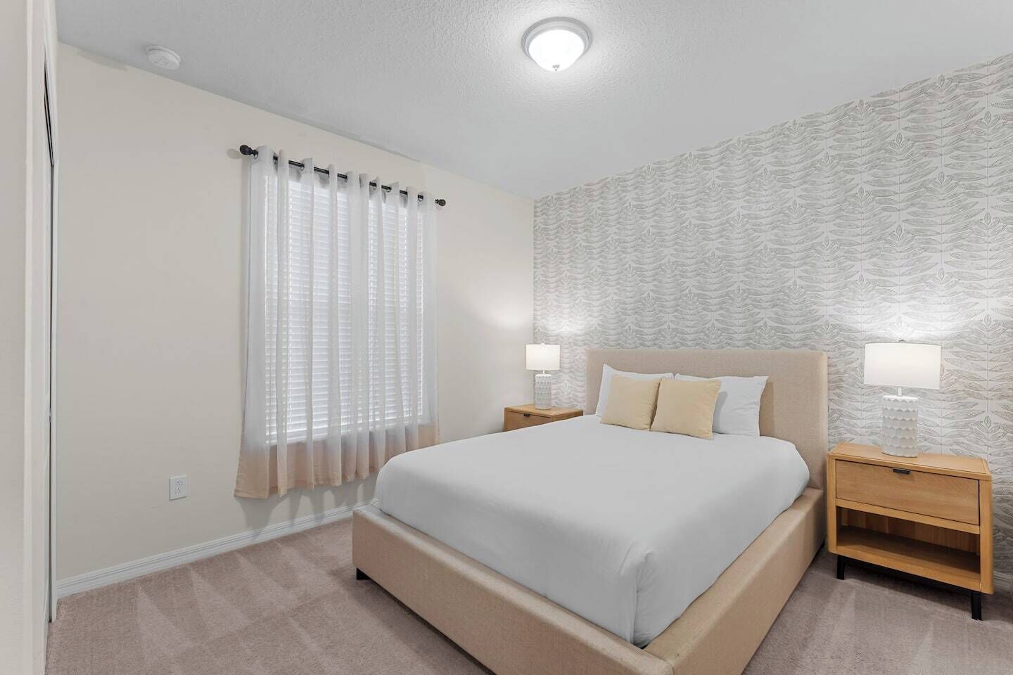A bedroom with a bed, two bedside tables with lamps, a window with sheer curtains, and a patterned wallpaper on the wall.