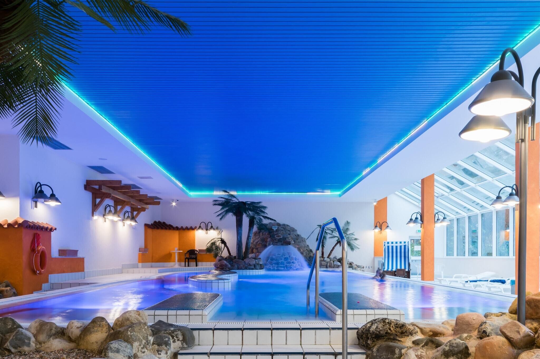 An indoor swimming pool with a waterfall feature, surrounded by rocks and palm trees, and equipped with modern lighting fixtures.