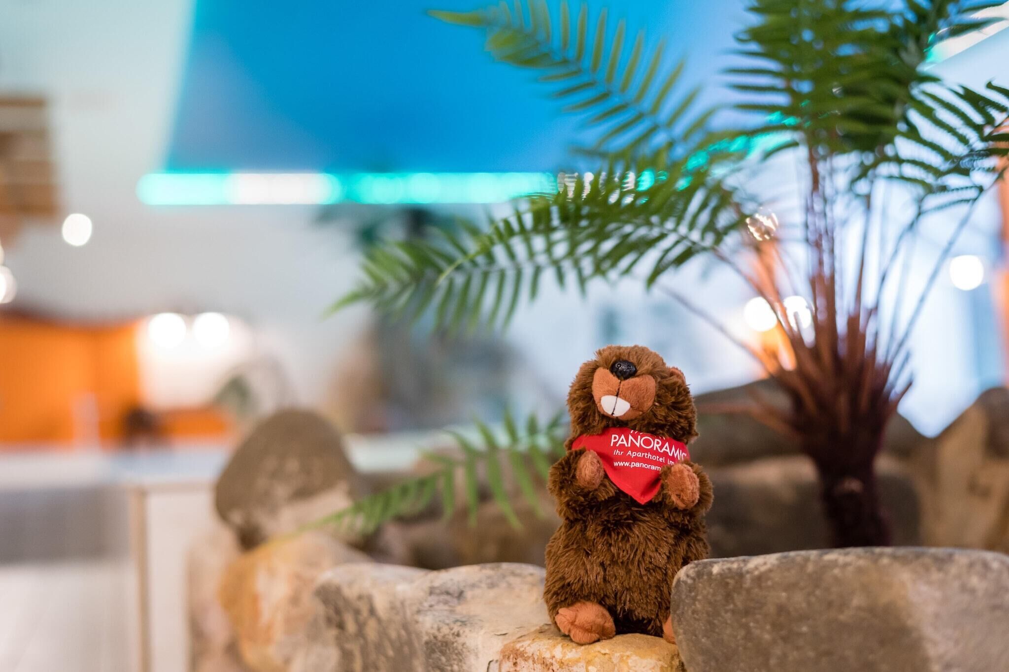 Un oso de peluche sobre una roca, con un lazo rojo, rodeado de plantas y un fondo desenfocado.