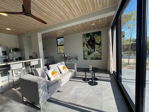 Ein moderner Wohnbereich mit grauem Sofa, Deckenventilator, Küche im Hintergrund und großem Fenster mit Blick ins Freie.