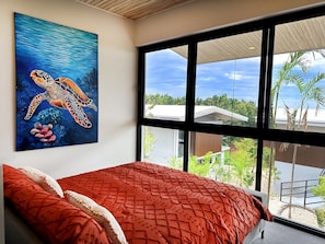 A bedroom with a large window, a red bedspread, and a painting of a sea turtle. - Solindo Stays (Cuajiniquil)