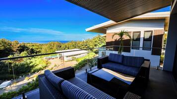 A balcony with outdoor seating, a view of the ocean, and a modern building in the background.