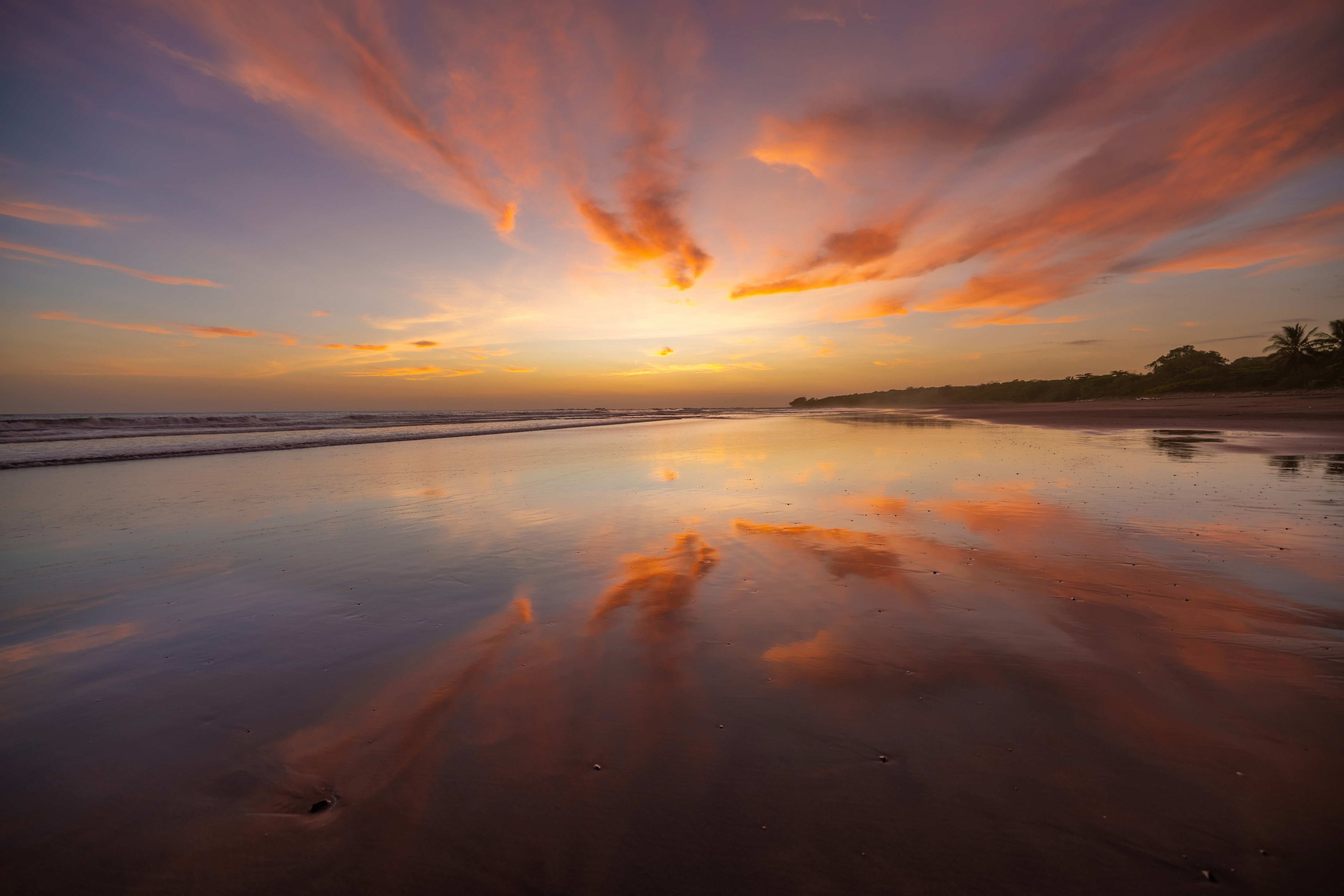 Uma praia com um pôr do sol, águas calmas e palmeiras ao longe.