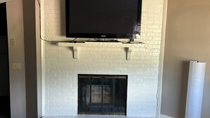 Una sala de estar con chimenea de ladrillo blanco, un televisor de pantalla plana montado y una lámpara de pie.