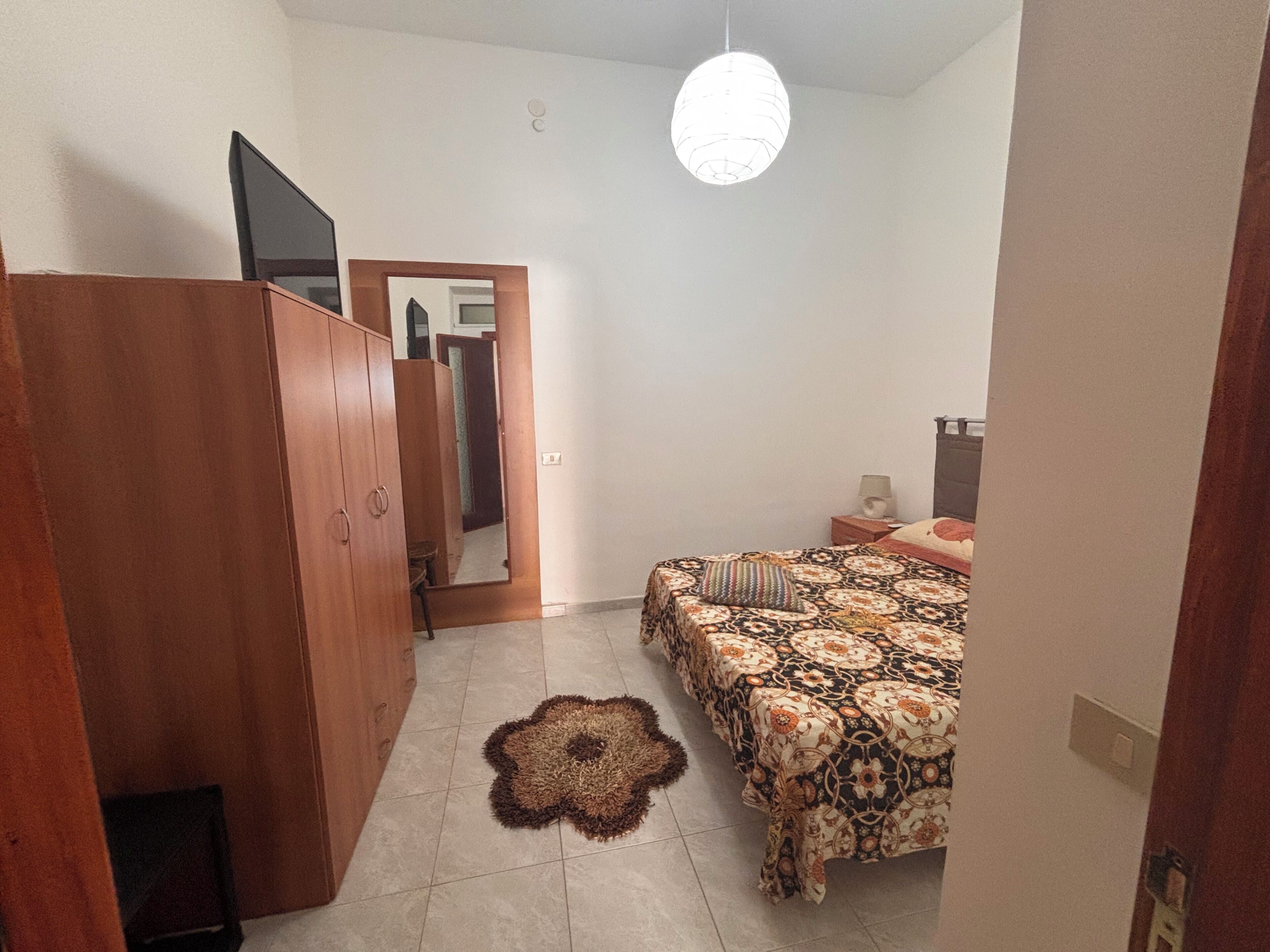 Ein Schlafzimmer mit Bett, Kleiderschrank, Spiegel, Teppich und einer Pendelleuchte.
