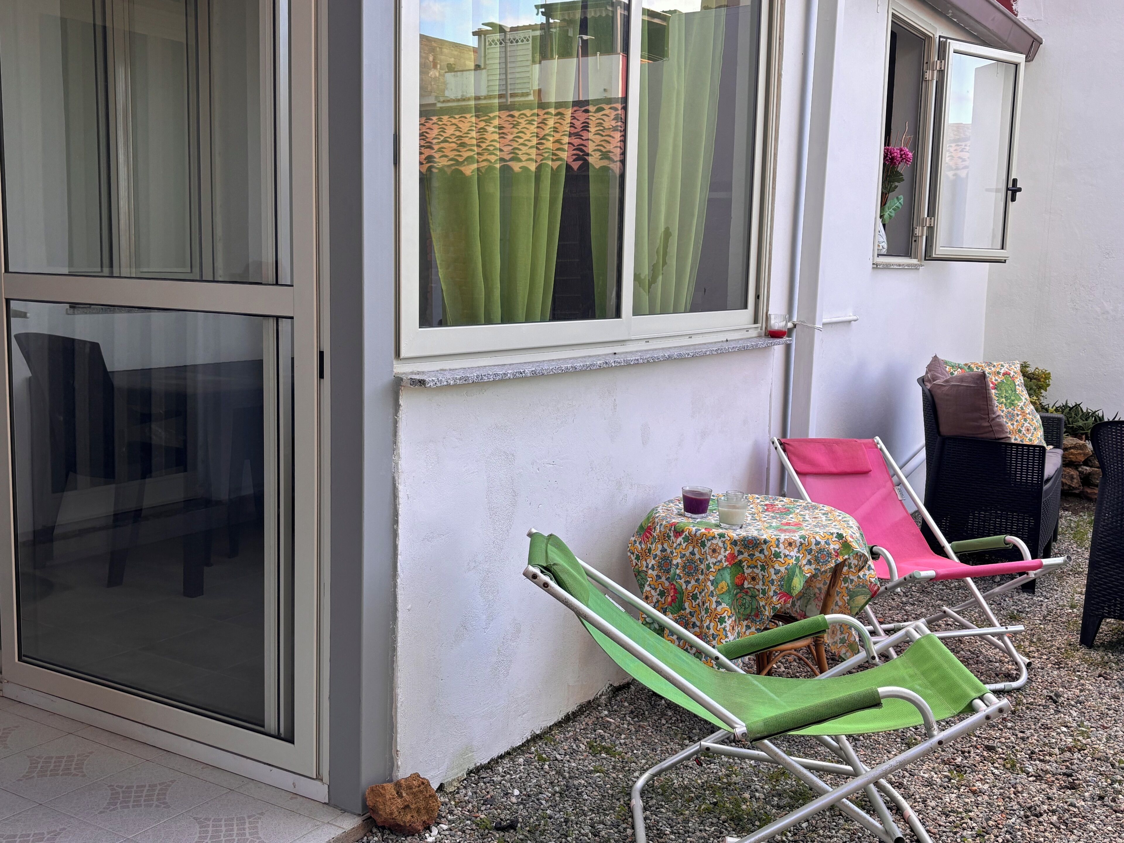 A room with a sliding glass door, a window with a curtain, a small table with a floral tablecloth, and two lounge chairs.