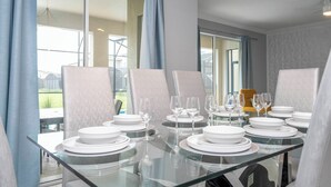 A dining table set with white plates and glasses, surrounded by cushioned chairs, in a room with large windows and a view of greenery.