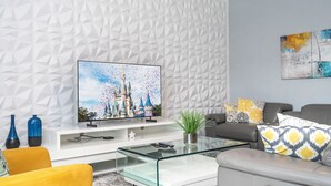 A modern living room with a geometric-patterned wall, a flat-screen TV, a glass coffee table, a sofa, and decorative pillows.