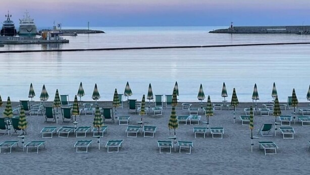 Une plage bordée de rangées de chaises de plage et de parasols aux rayures vertes et jaunes, une mer calme et, au loin, un navire amarré à un quai.