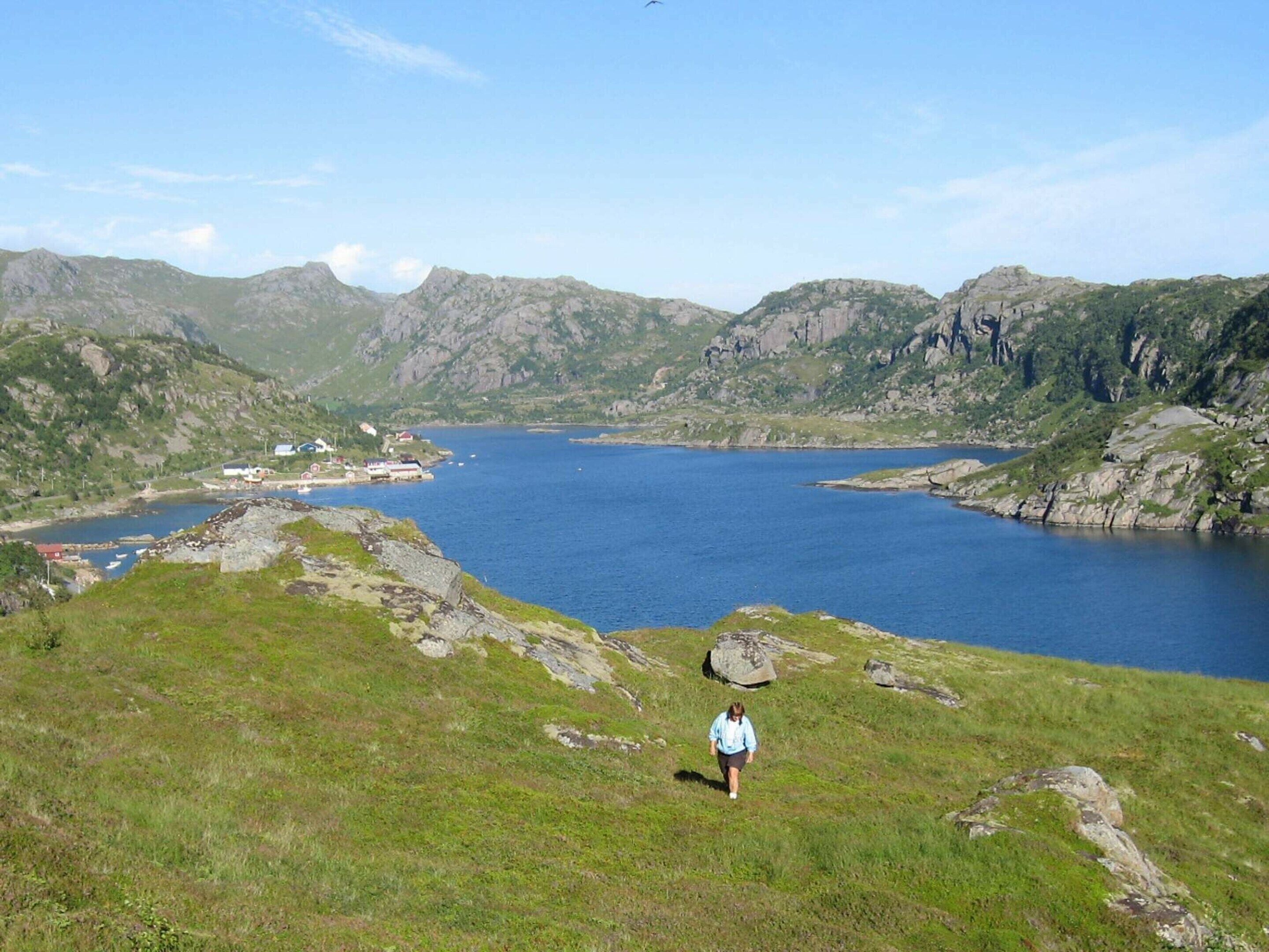 A person walking on a grassy hill overlooking a large body of water with mountains in the background.