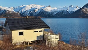 A small house with a dark roof and a balcony overlooking a calm lake with snow-capped mountains in the background. - 5 Person Holiday Home in Laukvik-by Traum (Vagan)