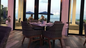 Un espace de restauration en plein air, avec des chaises en osier et une table dressée pour deux, donnant sur une plage parsemée de parasols.