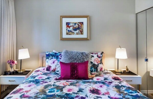 A hotel room with a floral bedspread, a framed artwork above the bed, and two bedside lamps.