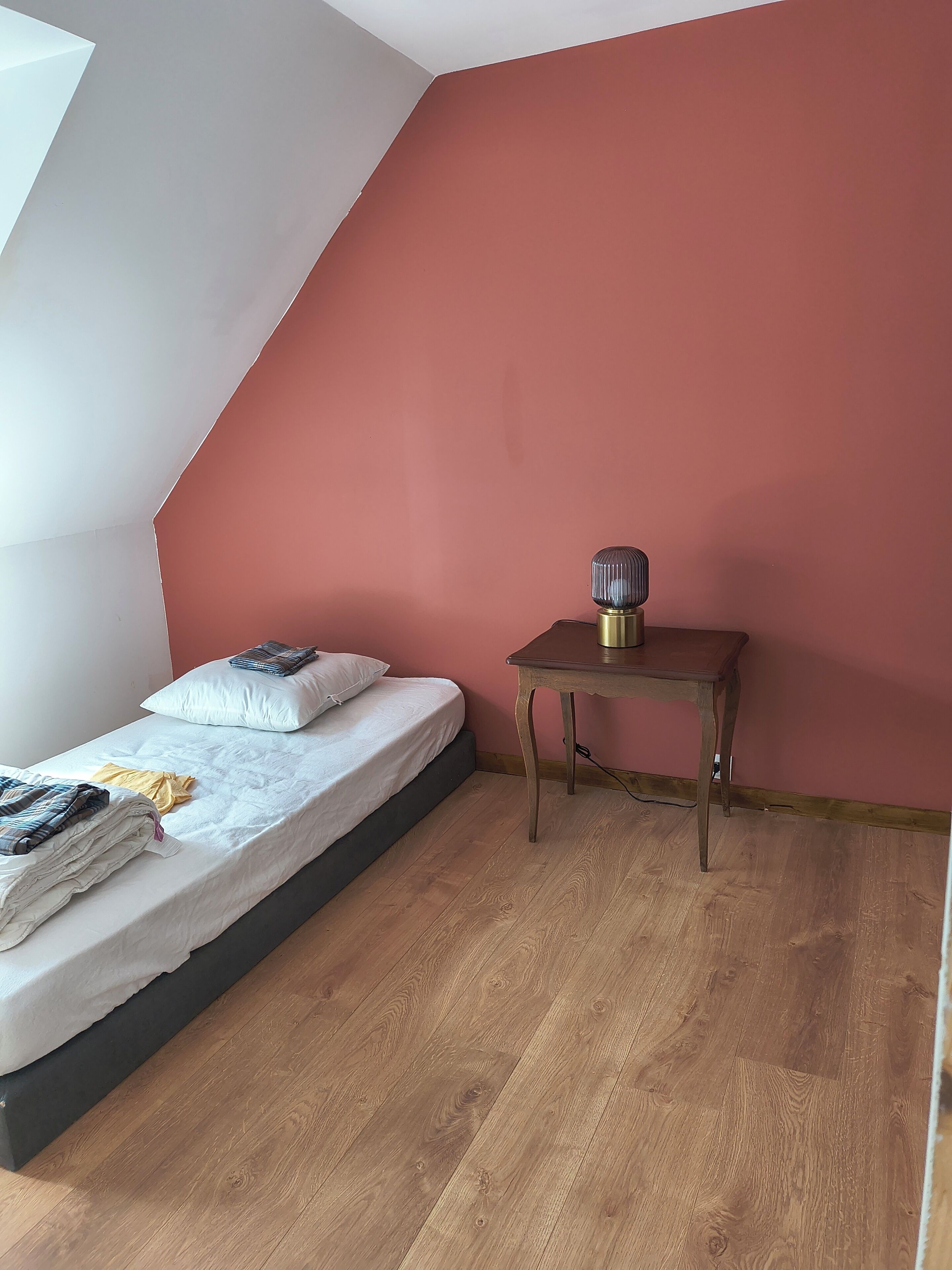 A bedroom with a sloped ceiling, a bed with pillows and a blanket, a small wooden table, and a red wall.