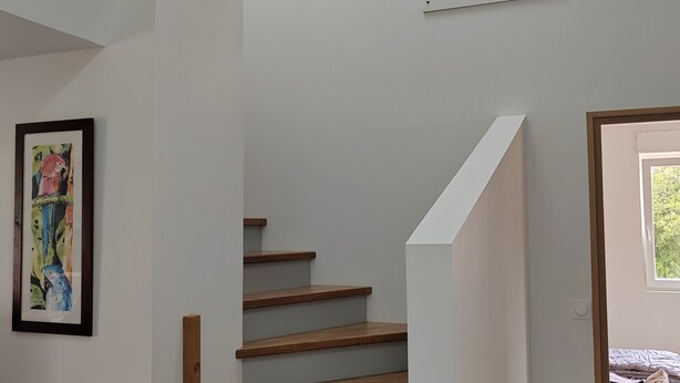 Un escalier avec des marches en bois et des murs blancs, un tableau encadré sur la gauche et une fenêtre sur la droite.