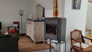 Un salon avec un poêle à bois, un canapé, un fauteuil, une table à manger et un escalier en arrière-plan.