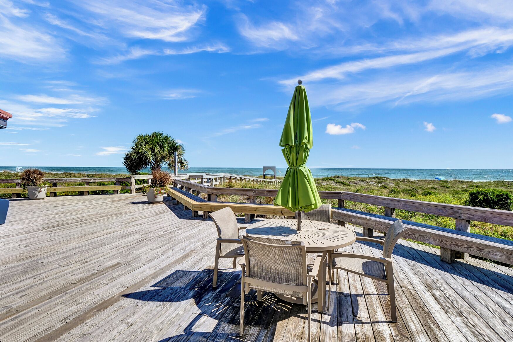 Une terrasse en bois avec des sièges d’extérieur et un parasol vert, offrant une vue sur une plage et l’océan.