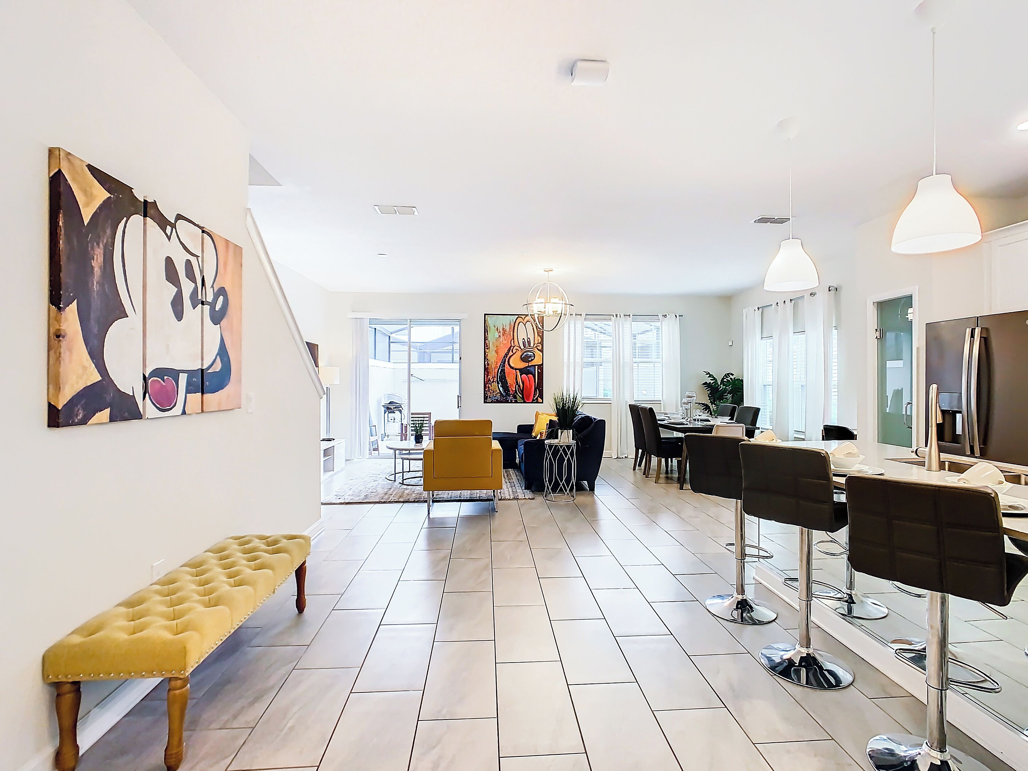 A modern living room with a bench, a dining area with chairs, a large painting, and a kitchen area with a refrigerator.