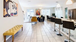 A modern living room with a bench, a dining area with chairs, a large painting, and a kitchen area with a refrigerator. - 5BR Townhouse Near Disney Pool (Kissimmee)
