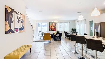 A modern living room with a bench, a dining area with chairs, a large painting, and a kitchen area with a refrigerator.