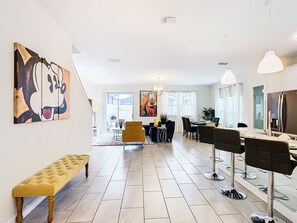 Uma sala de estar moderna com um banco, área de jantar com cadeiras, uma pintura grande e cozinha com geladeira.