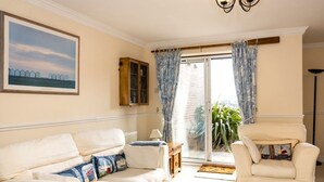 A living room with a white sofa, a matching armchair, a wooden cabinet, a painting, and a view through a glass door.