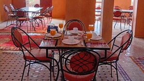 A dining area with a table set for four, surrounded by chairs with red cushions. The room has a tiled floor and arches, with a view of the outside. - Hotel Kasbah Riad Amnir (M'Hamid El Ghizlane)