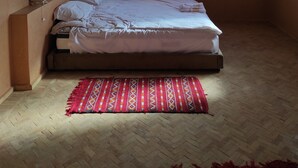 A bedroom with a wooden ceiling, a bed with white bedding, a red patterned rug, and a window with blinds. - Hotel Kasbah Riad Amnir (M'Hamid El Ghizlane)