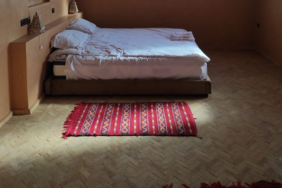 A bedroom with a wooden ceiling, a bed with white bedding, a red patterned rug, and a window with blinds.