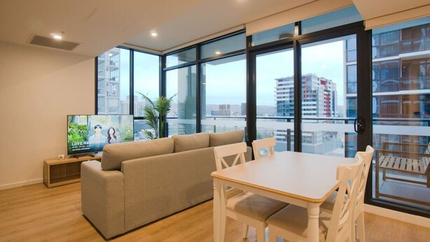 A modern living room with a sofa, dining table, and chairs. There is a TV on a stand, and large windows offer a view of the city. - City Serenity: 2BR Apt with Pool, Gym, Steam&Sauna (Adelaide)