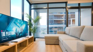 A modern living room with a large flat-screen TV displaying 'The Platform 2' poster, a beige sofa, wooden flooring, and large windows offering a view of a building with balconies. - City Serenity: 2BR Apt with Pool, Gym, Steam&Sauna (Adelaide)