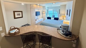 A hotel room with a curved desk, two wicker chairs, a bed, a TV, and a view of the sea. - Oceanfront Escape - Studio with kitchenette  (Ladysmith)