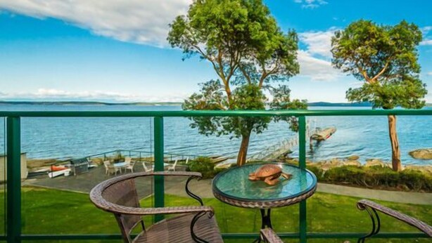 A terrace with a glass table and chairs overlooking a body of water, with trees and a view of the horizon. - Oceanfront Escape - Studio with kitchenette  (Ladysmith)