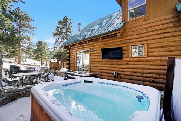 Una cabaña de madera con jacuzzi, mobiliario exterior y vistas a los árboles circundantes.