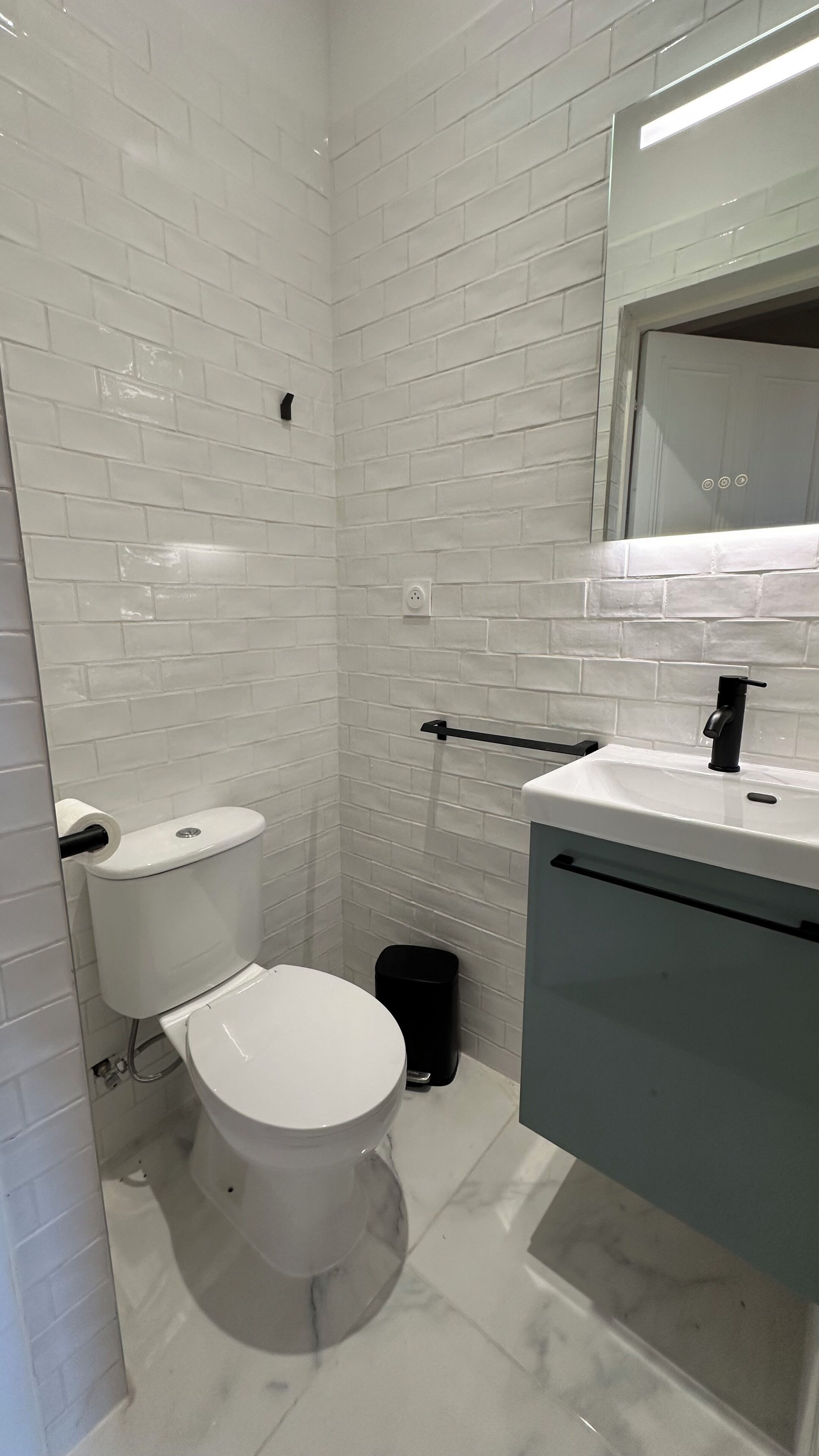 Une salle de bain avec des carreaux de céramique blancs rectangulaires, des toilettes blanches, une poubelle noire et un lavabo avec un robinet noir.