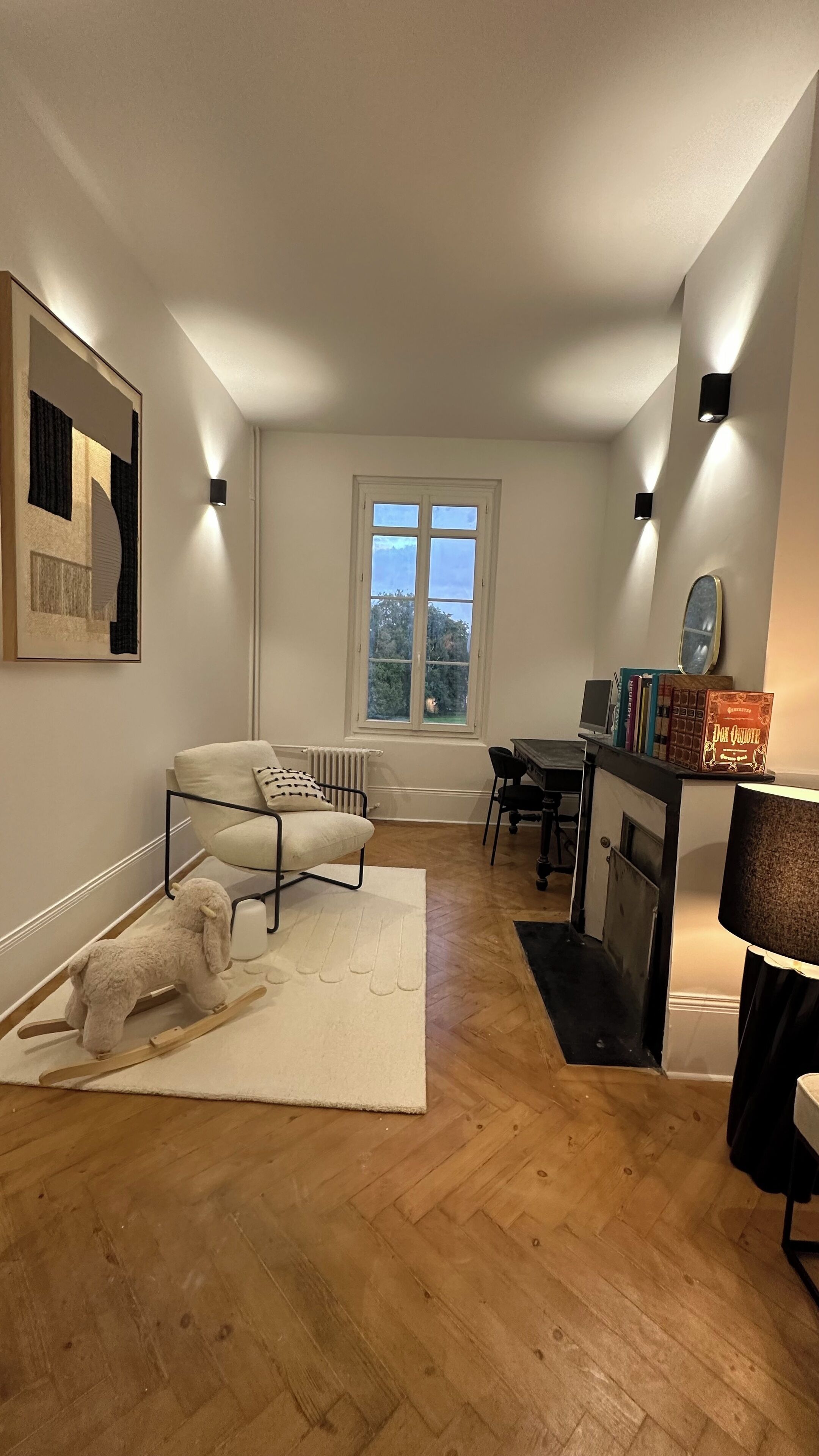 A modern living room with a sofa, a small table, a bookshelf, and a window with a view of trees.