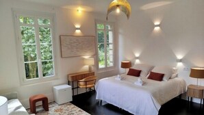 A bedroom with a large bed, bedside tables, a desk, and chairs. There are windows with views of greenery. - Chateau Loyd&Spa Normandie Pays de Caux (Les Hauts-de-Caux)