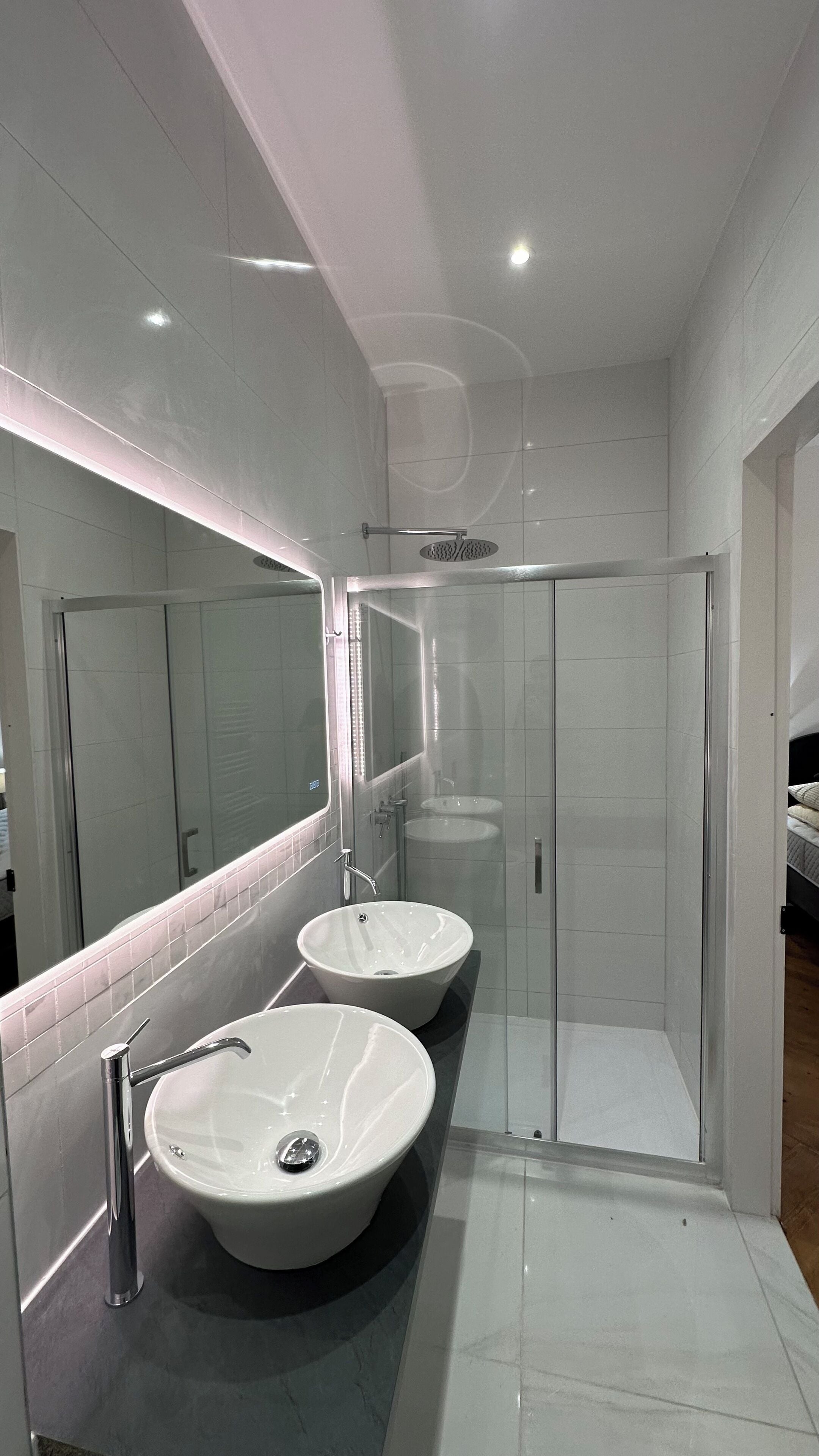 Un baño moderno con dos lavamanos grandes de color blanco, cabina de ducha de cristal e iluminación LED en el espejo.