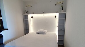 A small, neatly made bed in a room with a white ceiling, a small shelf with plants, and a simple light fixture.