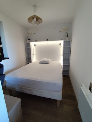 A small, neatly made bed in a room with a white ceiling, a small shelf with plants, and a simple light fixture.