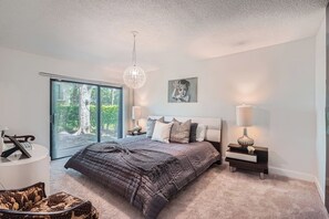 A bedroom with a large bed, two bedside tables, a chandelier, and a view of greenery through a sliding glass door.