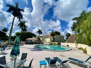 A pool area with lounge chairs and umbrellas, surrounded by palm trees and buildings.