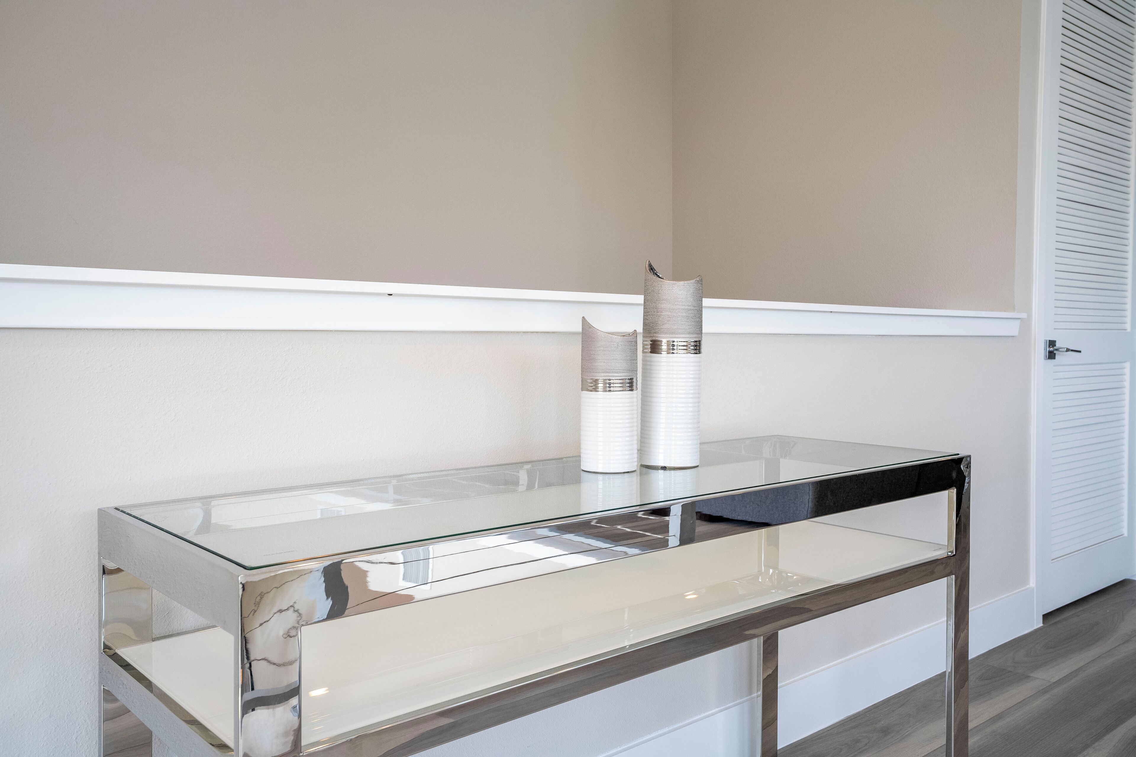 Une table à plateau en verre et à structure métallique, placée contre un mur avec un plinthe blanche et une porte dotée de volets à persiennes.