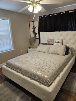 A bedroom with a tufted headboard, a bed with a patterned bedspread, a bedside table with a lamp, a clock, and a framed picture on the wall.