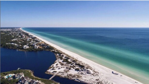 Luftaufnahme einer Küstenlandschaft mit einem langen Sandstrand, einem Yachthafen und Wohngebäuden.