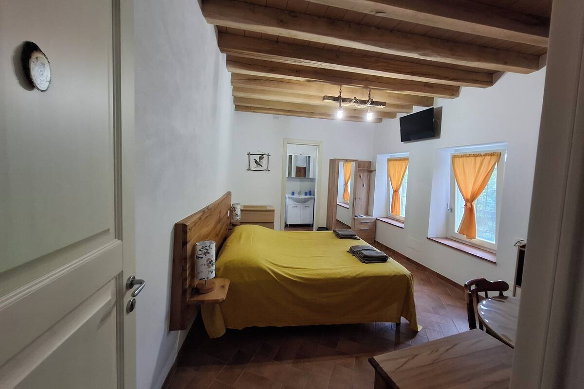A bedroom with a wooden bed, a bedside table, a mirror, a window with curtains, and a wooden ceiling.