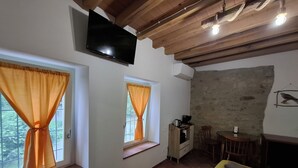 A room with a TV mounted on the wall, two windows with curtains, a sofa, a small table, and a stone wall. - Il Castellaro ai Sassi (Guiglia)