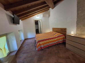 A bedroom with a wooden ceiling, a bed with a striped blanket, a bedside table with a lamp, and a TV mounted on the wall. - Il Castellaro ai Sassi (Guiglia)