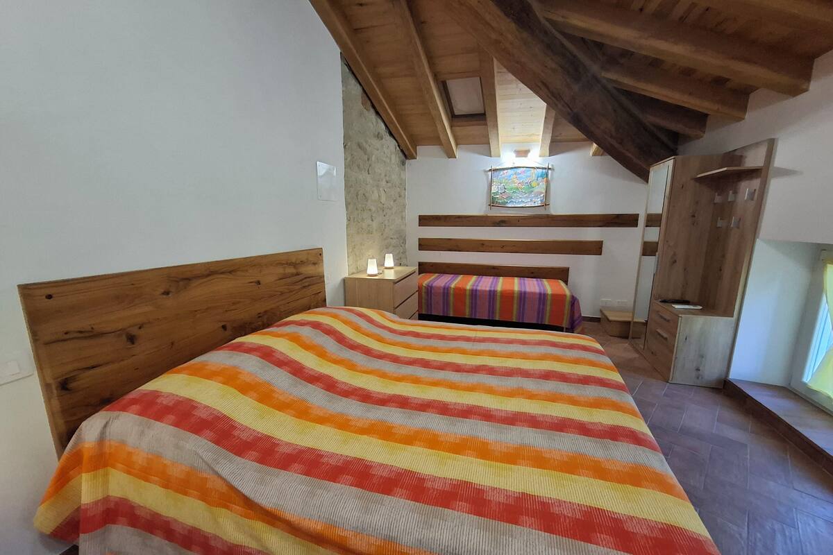A bedroom with a wooden ceiling, a bed with a striped blanket, a nightstand with lamps, and a small window with a painting.