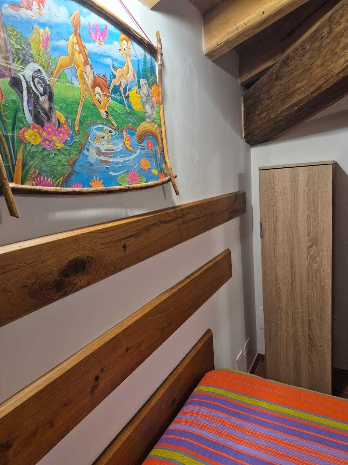 Een slaapkamer met een houten hoofdeinde, een kleurrijke strepenbedekking, een schilderij van tekenfiguren aan de muur en een houten garderobe.